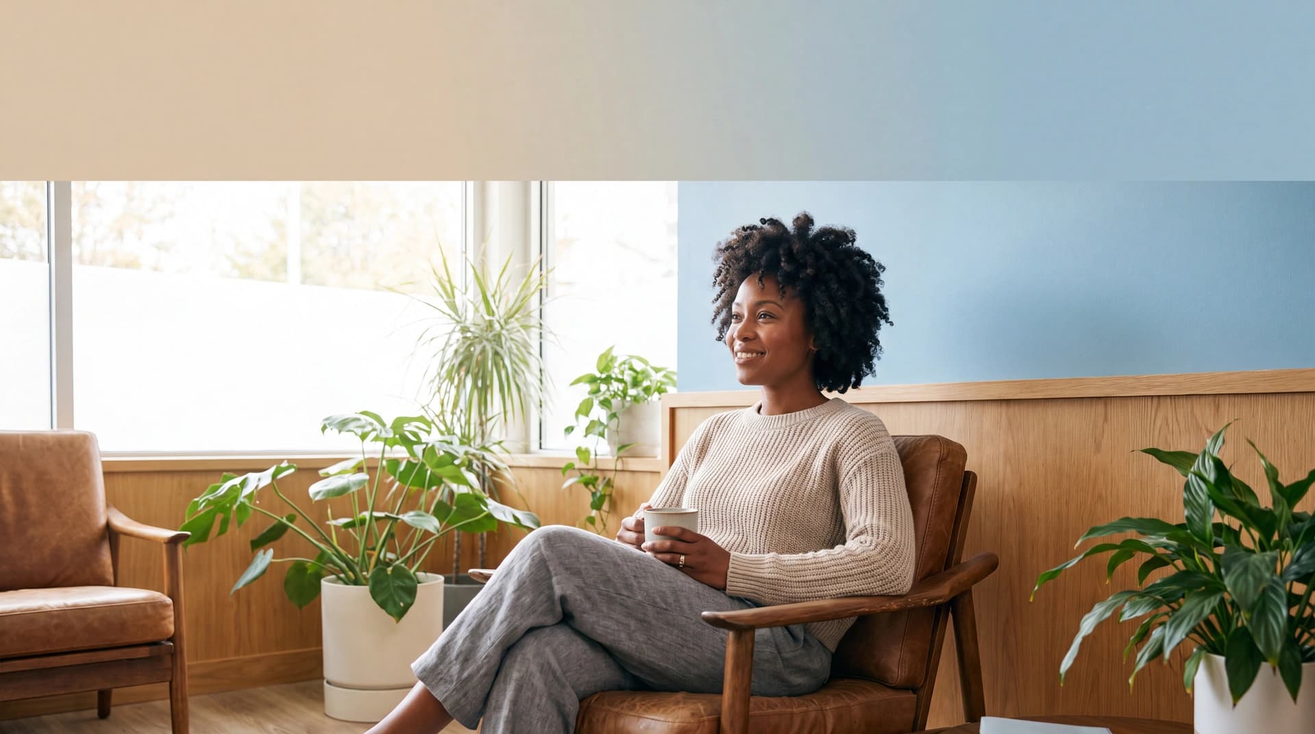 A woman sits comfortably in a warm, plant-filled medical waiting area with soft daylight